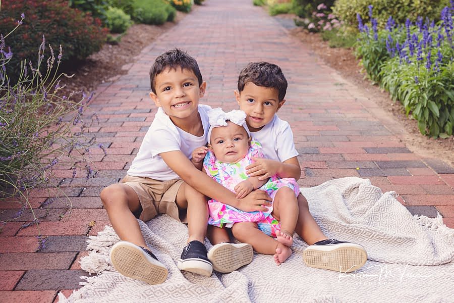 Older brothers sitting on a blanket holding their baby sister during their photoshoot by The Flash Lady Photography