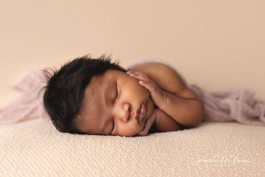 Sleeping black baby girl holding her cheeks posing on a cream blanket during her photoshoot by The Flash Lady