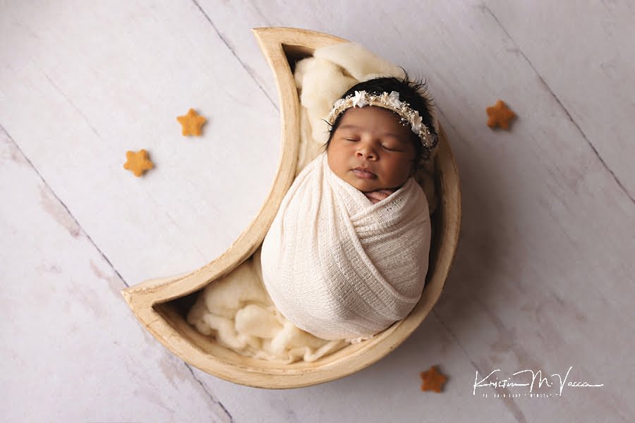 Sleeping black baby girl wrapped in white posing in a moon prop on a white wood background during her photoshoot