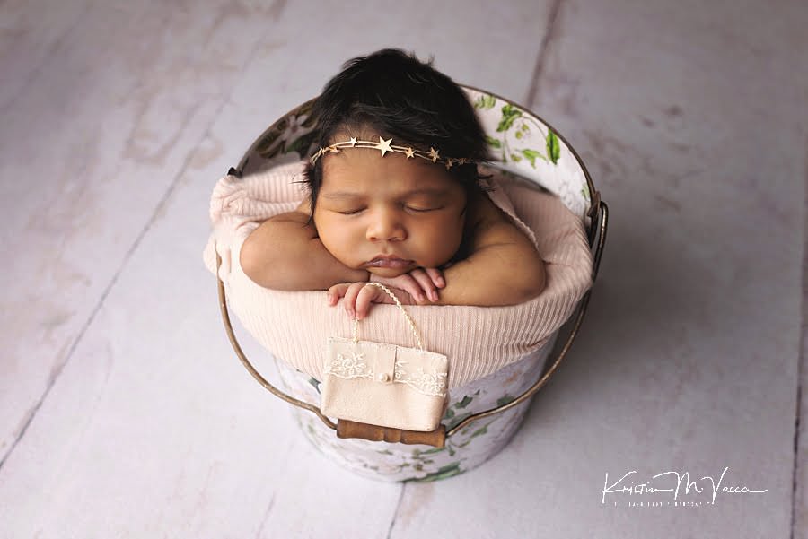 Black baby girl posing in a floral bucket holding a pink purse during her beautiful newborn photography session by The Flash Lady
