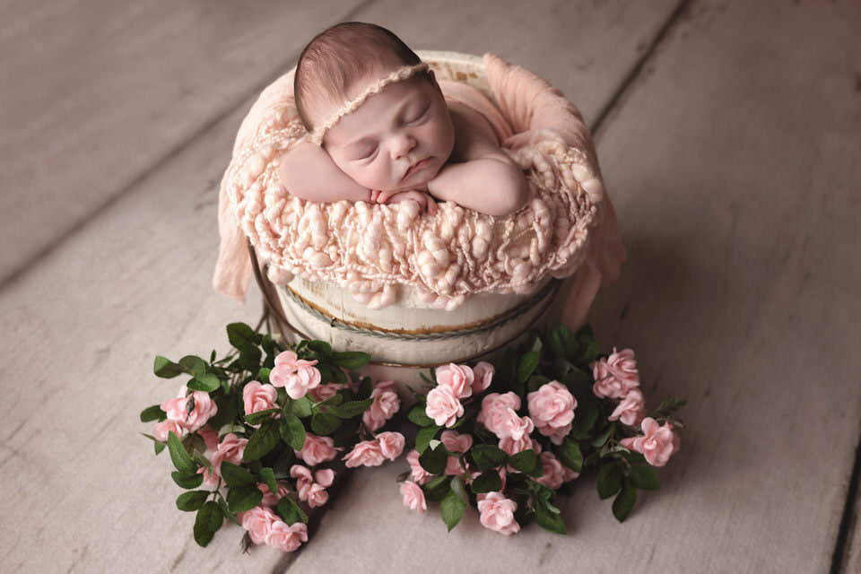 Sleeping newborn baby girl lying over a pink blanket in a white bucket surrounded with pink roses during her newborn photoshoot