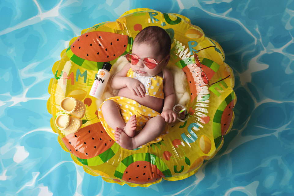 Sleeping newborn baby girl with pink heart sunglasses in a yellow summer patterned pool float during her photoshoot