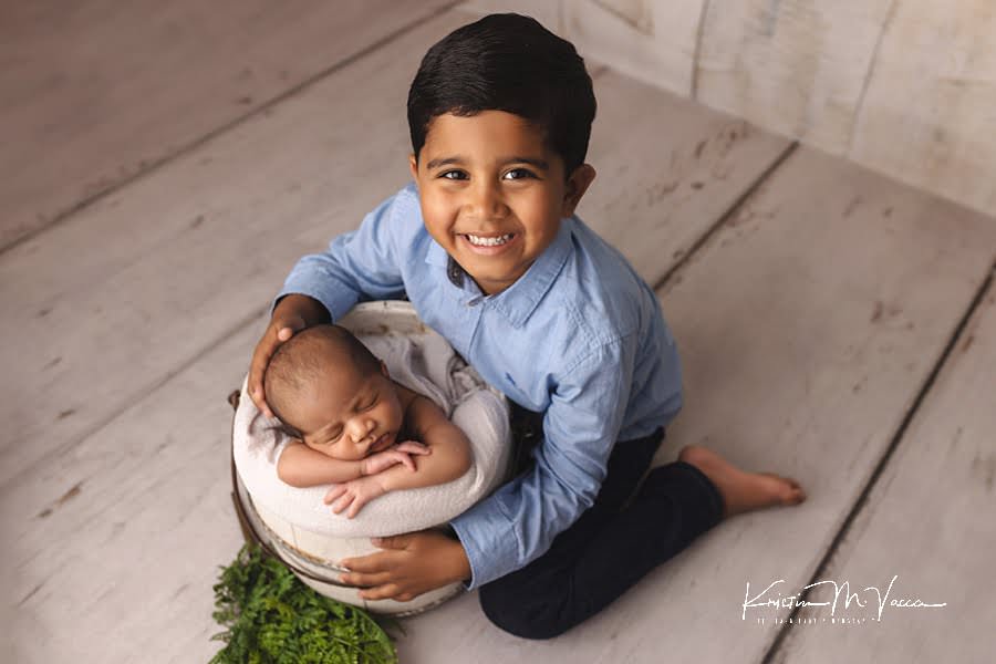 Indian big brother smiling at the camera as his sleeping newborn brother posing in a white bucket during their photoshoot