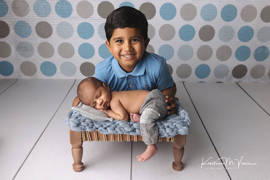 Indian newborn baby boy sleeping on a blue blanket and brown platform while his big brother smiles at the camera during their newborn photos with older brother