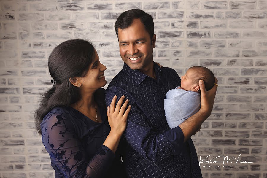 Husband and wife from India smiling as each other as they hold their newborn baby boy wrapped in blue during their photography session
