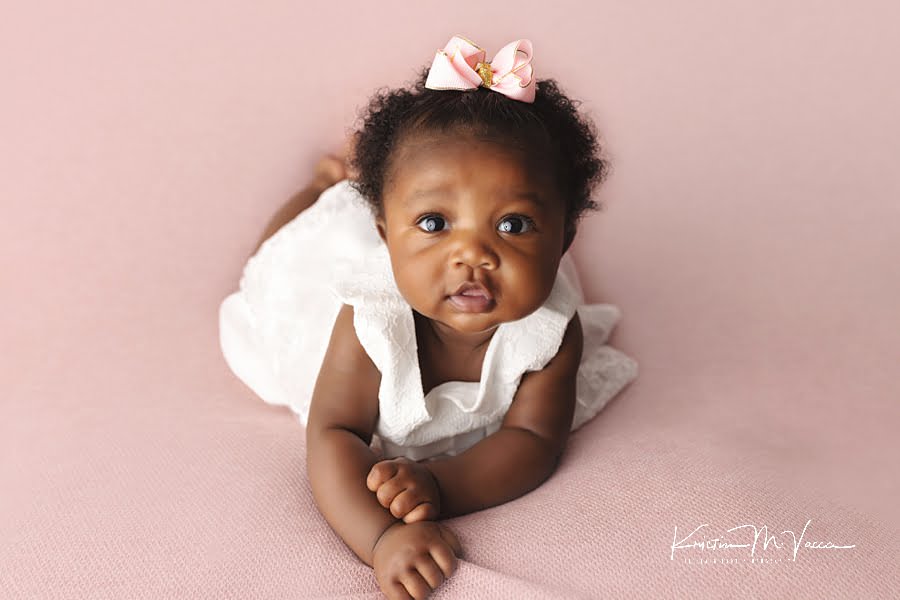 Black baby girl in a white dress posing on a pink blanket during her milestone photoshoot