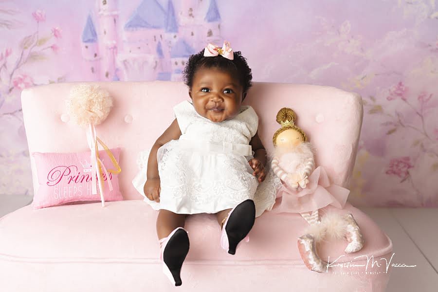Smiling black baby girl posing in a white dress and heels on a pink couch during her princess baby photos by The Flash Lady