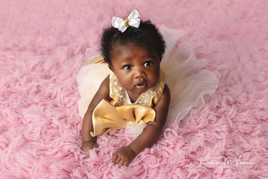 Black baby girl lying on her stomach on pink fur in a gold dress during her princess baby photos by The Flash Lady