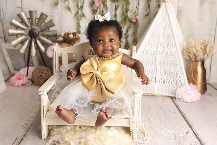 Smiling black baby girl sitting on a cream bench in front of a bohemian theme background during her photoshoot by The Flash Lady