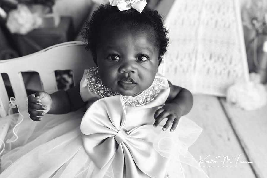 Black and white photo of a black baby girl looking at the camera sitting on a white bench during her photoshoot