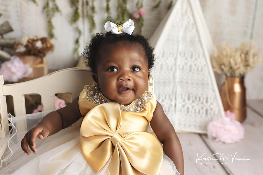 Smiling black baby girl in a gold dress sitting on a cream bench posing during her princes baby photos by The Flash Lady