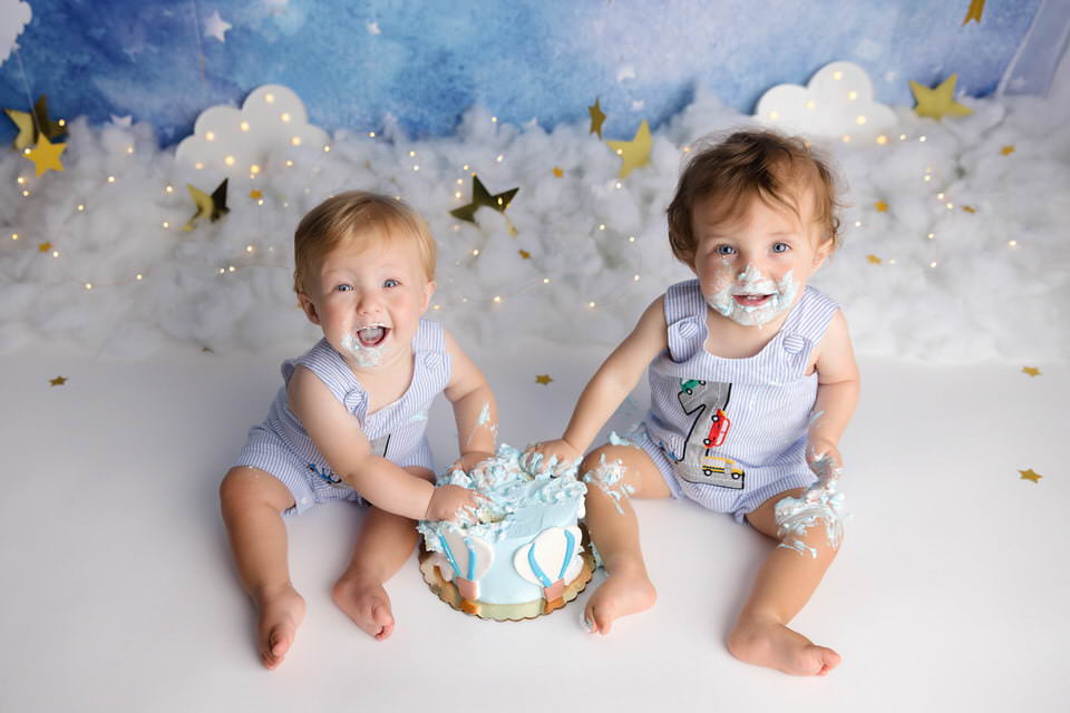 Smiling twin boys looking up at the camera as they smash their birthday cake in front of a black background during their cake smash photoshoot