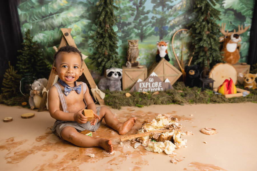Smiling black toddler smiling as he smashes his cake in front of a camping theme during his first birthday photoshoot by The Flash Lady