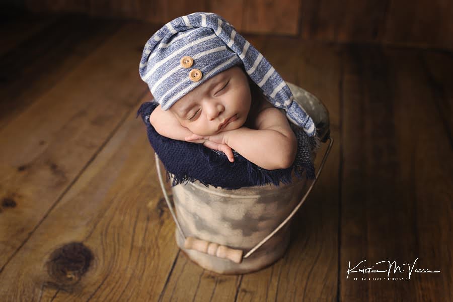 Baby boy sleeping in a bucket in a blue and white striped hat during his photoshoot