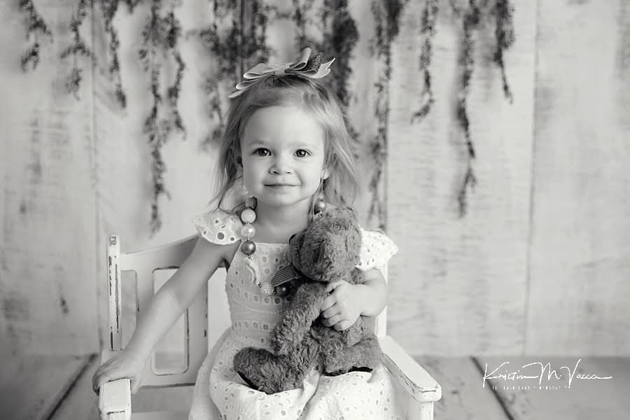 Black and white photo of a toddler girl holding a teddy bear sitting in a chair during her paint smash photoshoot