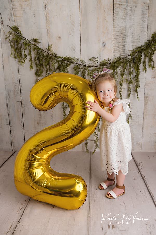 Toddler girl smiling and holding a gold #2 balloon during her photoshoot