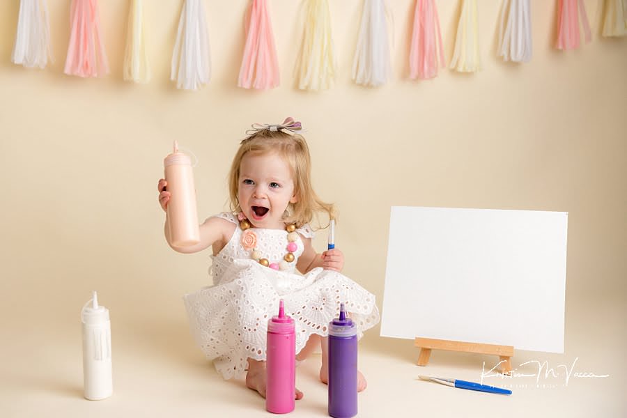 Excited toddler girl holding a peach paint bottle in amazement during her paint smash photoshoot