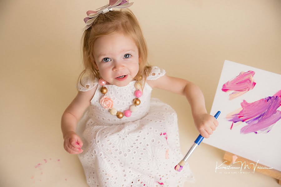 Smiling toddler girl looking up at the camera holding a brush during her paint smash photoshoot