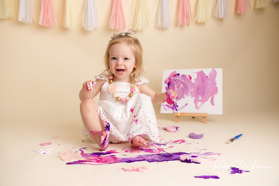 Toddler girl laughs as her feet at in pink and purple paint during her paint smash photoshoot