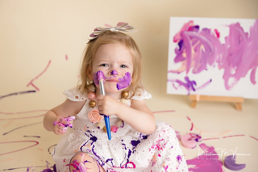 Toddler girl paints her cheeks with purple paint brush during her photoshoot