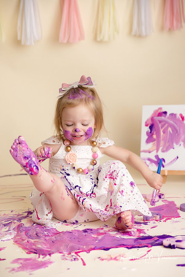 Toddler girl smiling and holding up her foot cover in paint during her photoshoot