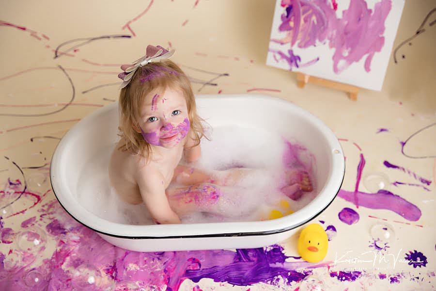 Photo from above of a toddler girl sitting in a bathtub after her paint smash photoshoot