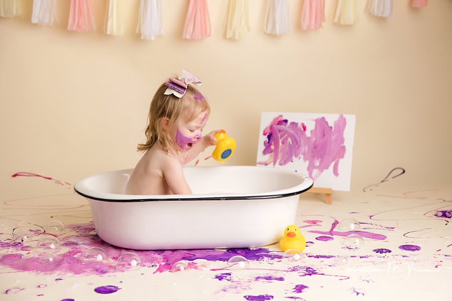 Toddler girl laughs while splashing in the tub after her paint smash photoshoot