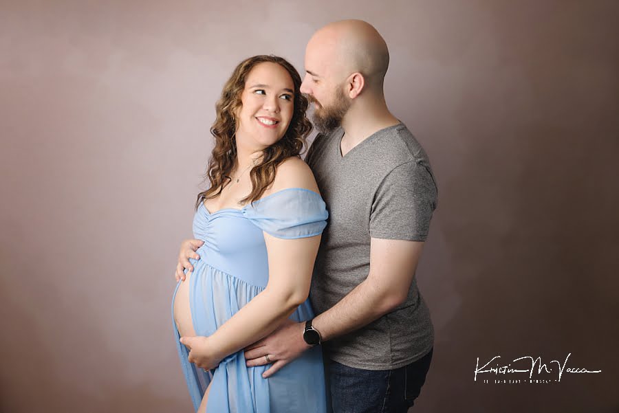 Husband standing behind his wife holding her belly while they look at each other during their beautiful studio maternity photos