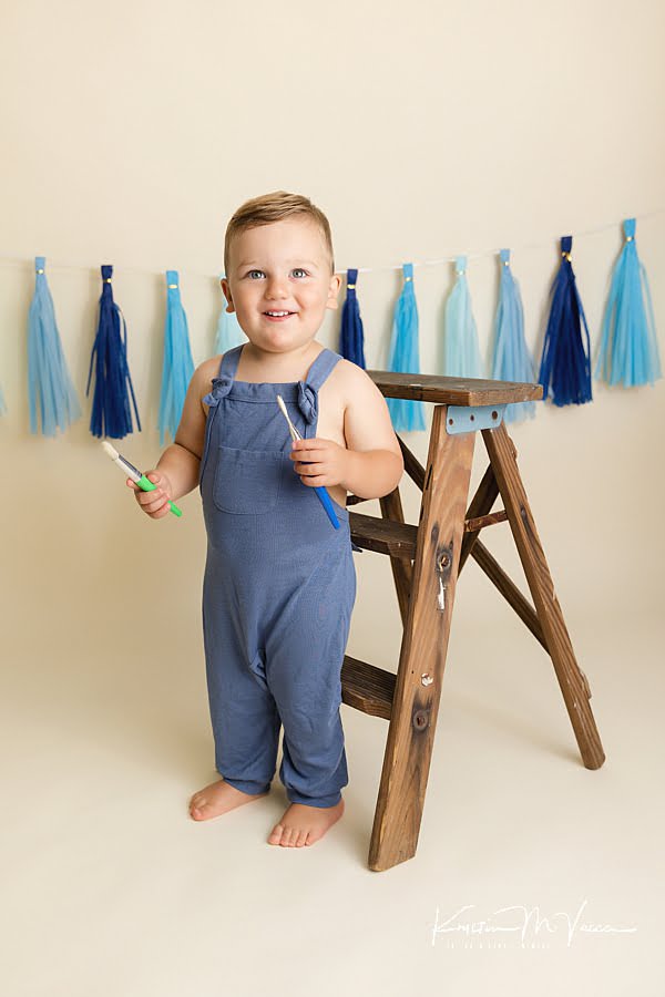 Smiling toddler boy standing in front of a ladder holding brushes during his blue paint smash
