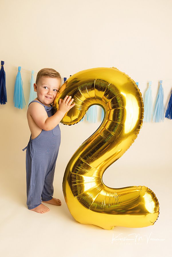 Smiling toddler boy holding a gold #2 large balloon during his birthday paint smash