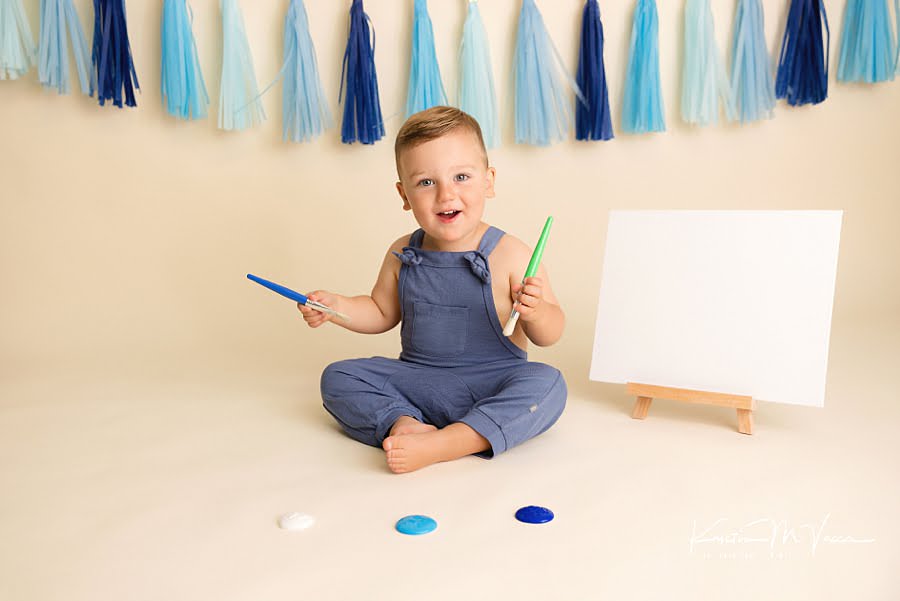 Toddler boy holding paint brushes and excited to paint during his birthday photoshoot