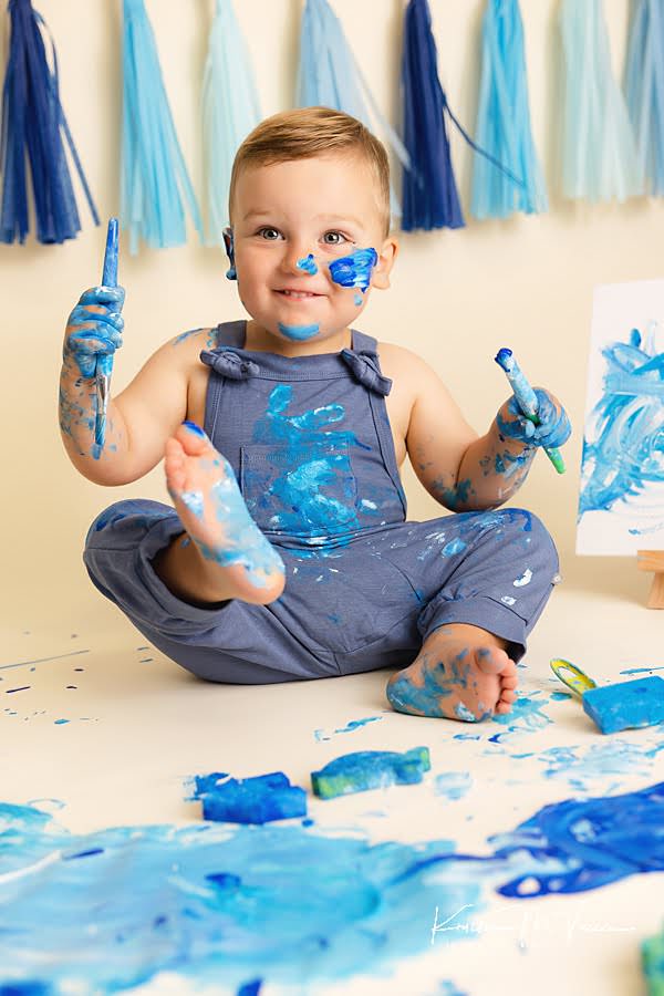 Smiling toddler boy holds his painted foot in the air during his blue paint smash