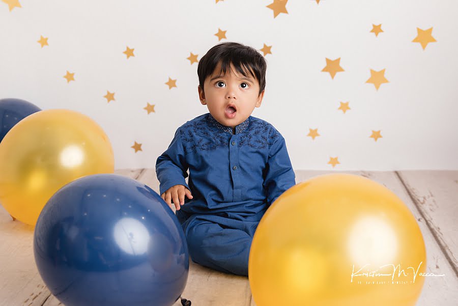 Toddler boy in blue outfit amazed at the balloons surrounding him during his cake smash photoshoot