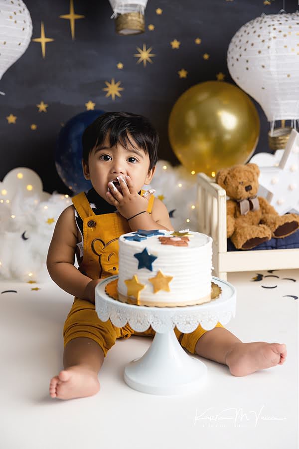 Toddler boy holds his hand up to his mouth to eat cake during his first birthday photoshoot
