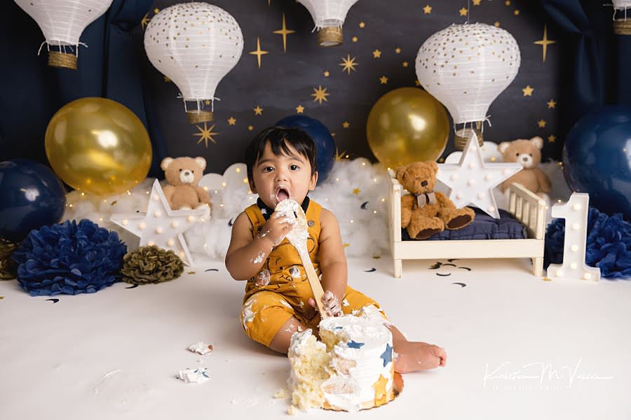 Toddler boy eats from a wooden spoon during his blue & gold teddy cake smash
