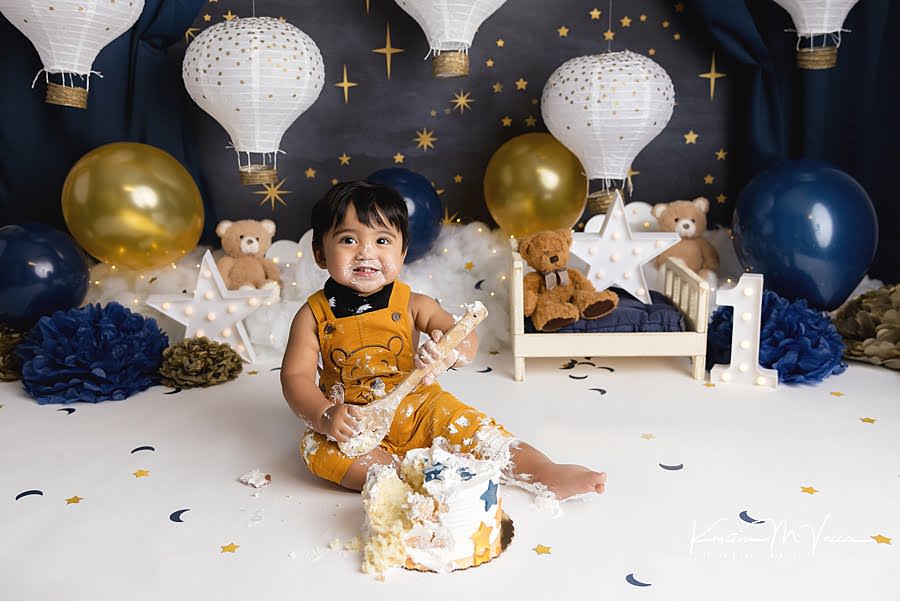 Toddler boy smiles as he's holding a wooden spoon during his blue & gold teddy cake smash