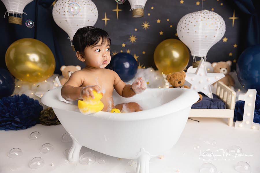 Toddler boy watches bubbles fall while sitting in a bathtub during his blue & gold teddy cake smash