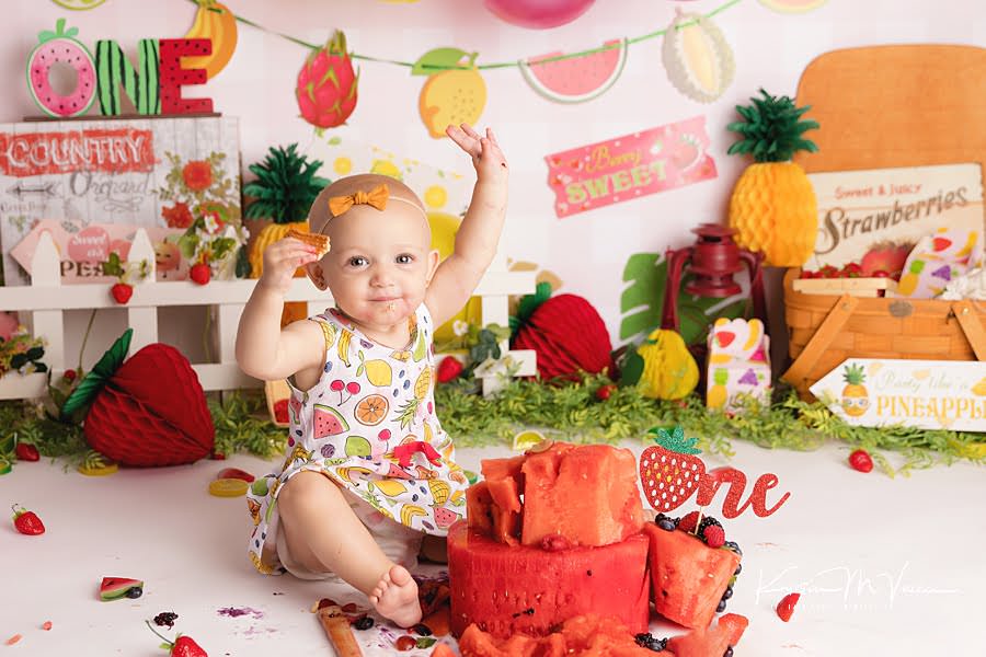 Baby girl holds her hands in the air while eating during her fruit cake smash