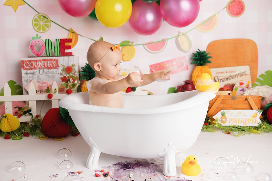 Baby girl points towards a rubber duck while taking a bath during her birthday photoshoot