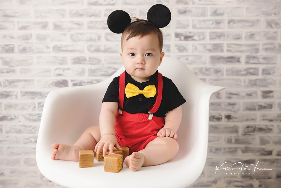 Baby boy sits in a white chair with blocks during his mickey carnival cake smash