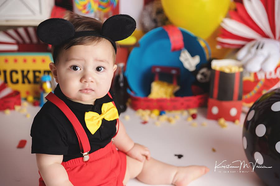 Baby boy sits in front of his mickey carnival cake smash scene for his birthday photos