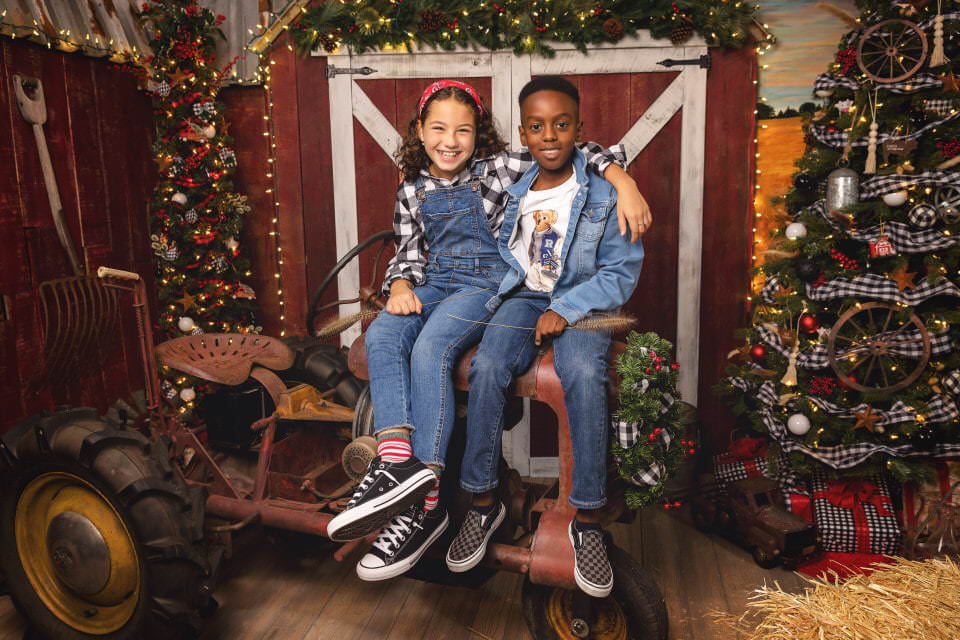 A young white girl and a black boy in denim sit on an antique tractor together during their Christmas photoshoot