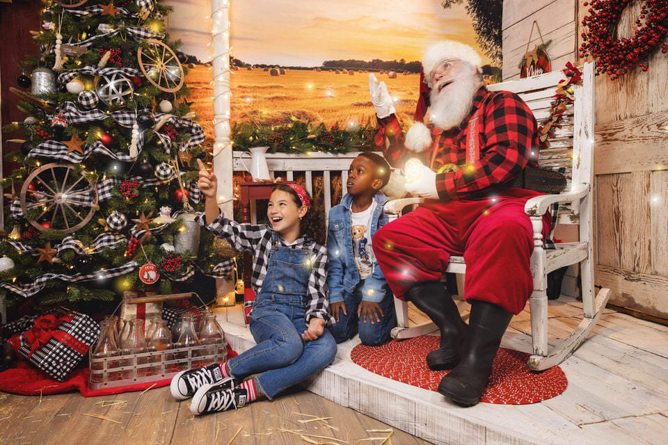 A young girl and boy sit on a porch step with Santa Claus amazed at the fireflies during a Christmas photoshoot