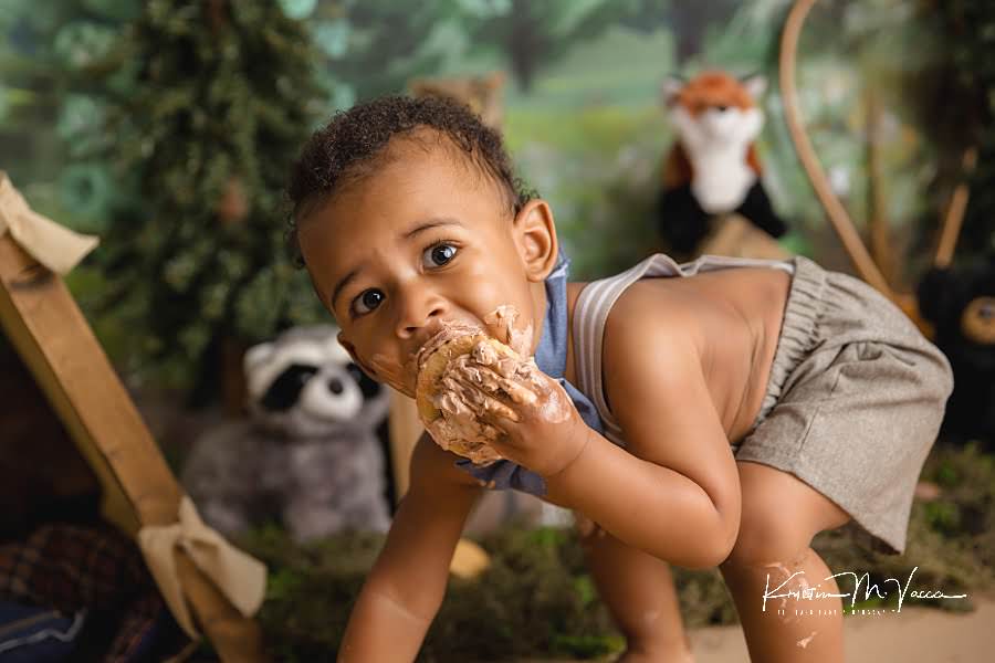 Toddler boy leans over while stuffing a frosted cookie in his mouth during his camping woodland cake smash