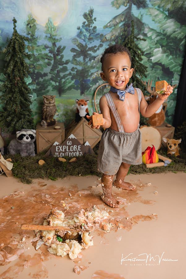 Smiling toddler boy holding 2 blocks behind his smashed cake during his camping woodland cake smash