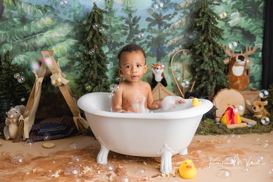 Toddler boy sitting in a bath tub surrounded by bubble during his camping woodland cake smash