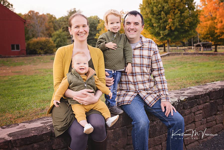Family of 4 sits on a rock wall smiling during their happy fall family photos