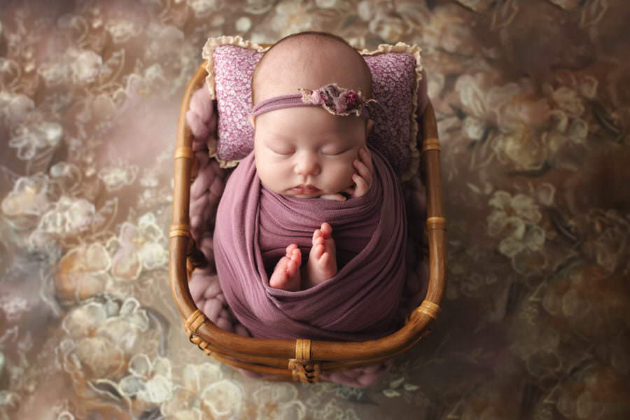 Sleeping newborn baby girl wrapped in purple posed in a basket during her photoshoot