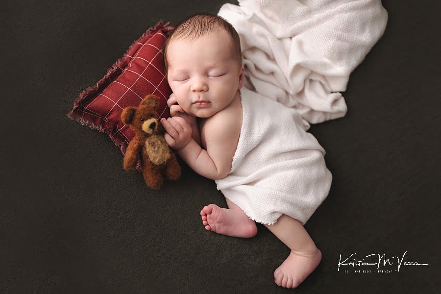 Sleeping baby boy curls up on a red pillow with his teddy bear during his gifted newborn photos