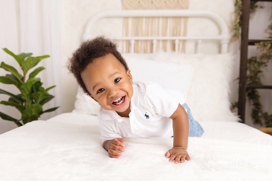 Black baby boy lies on a bed smiling during his 1st birthday milestone photos